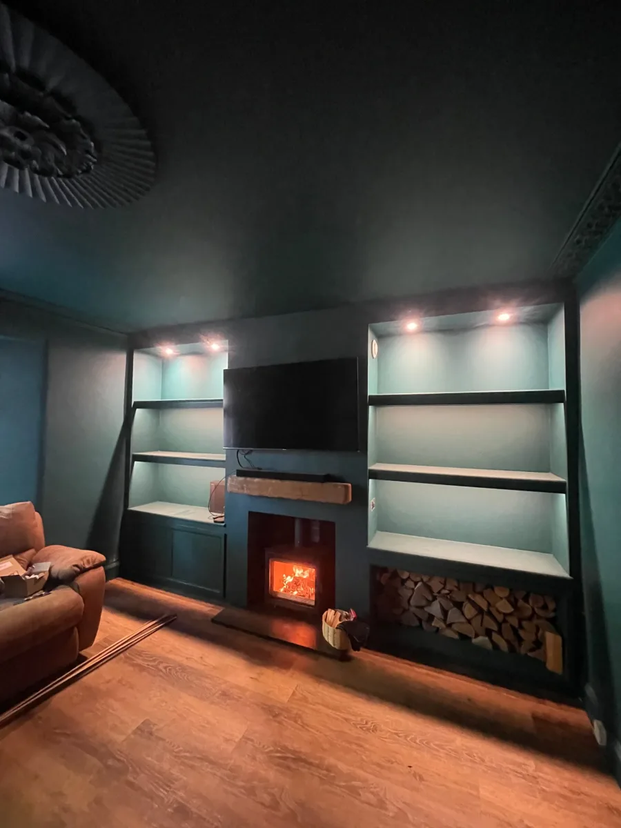 Wood burning stove installed in dark teal living room with built-in shelving log store and TV above fireplace