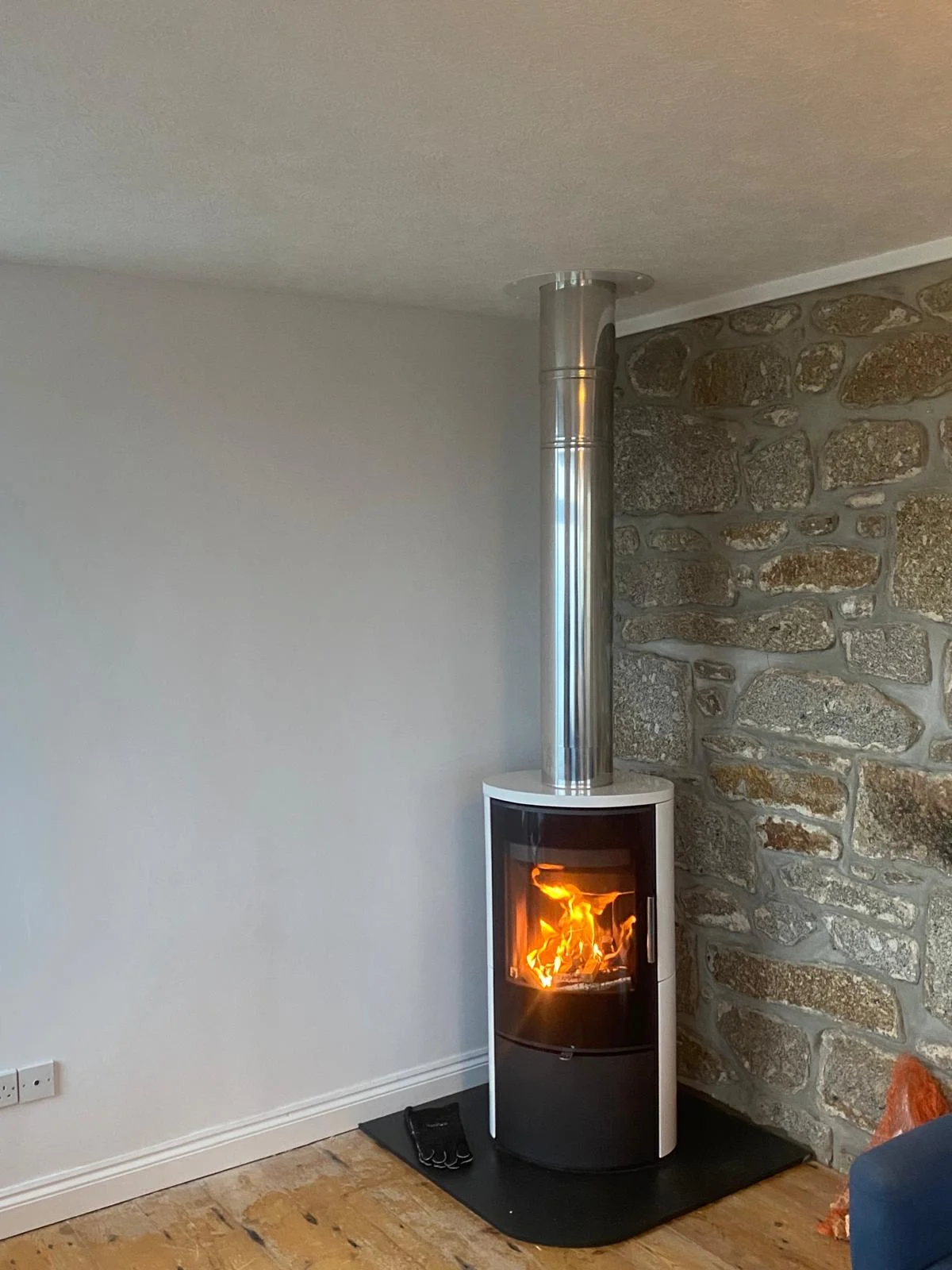 Modern cylindrical wood burning stove black white with twin wall flue pipe installed against Cornish stone wall