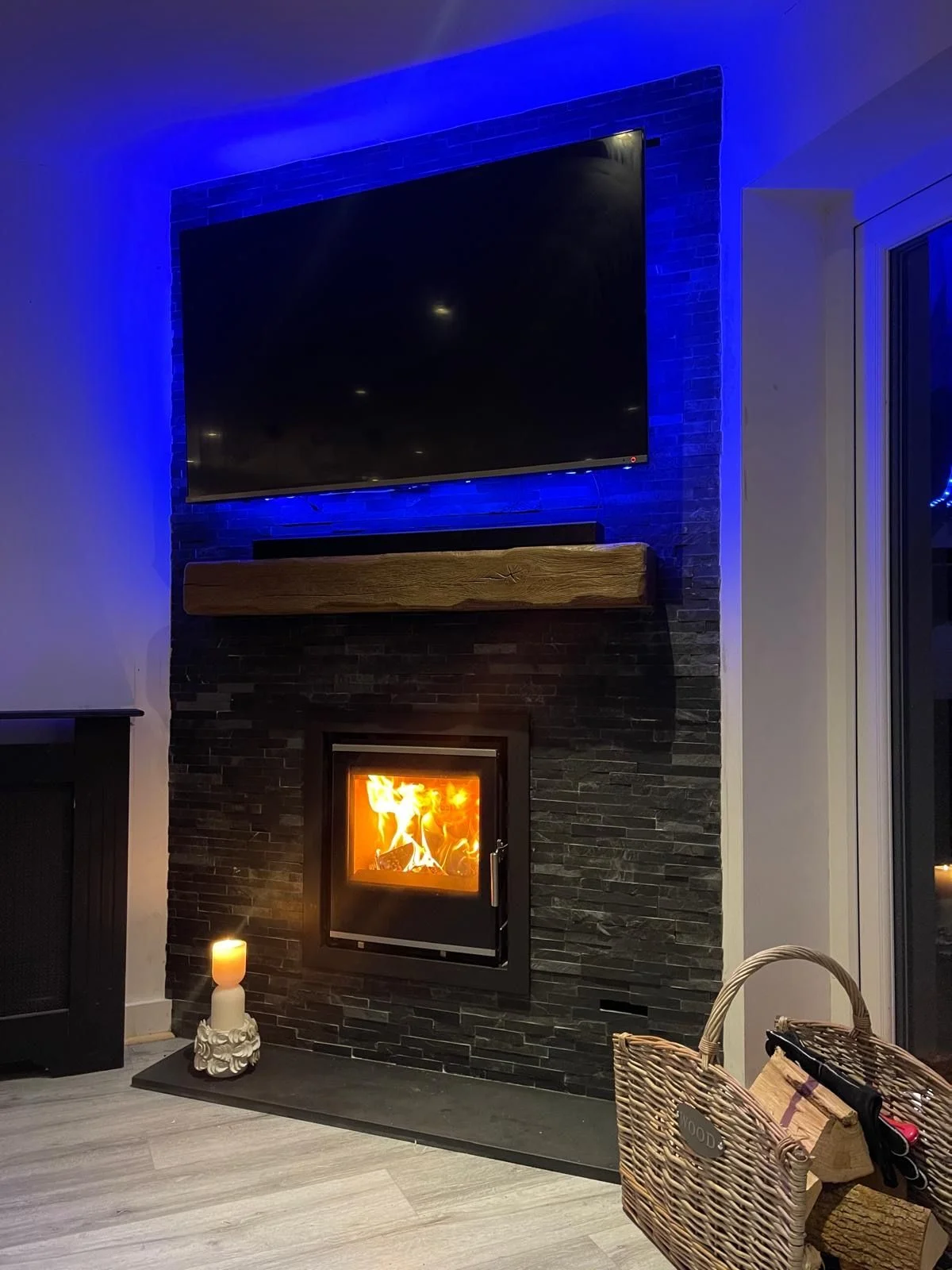 Inset wood burning stove in slate wall with oak beam mantel blue LED lighting and wall-mounted TV