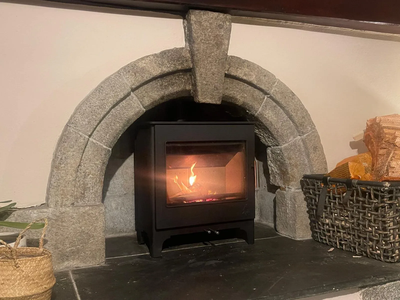 Black wood burning stove alight set within traditional Cornish granite stone arch fireplace slate hearth