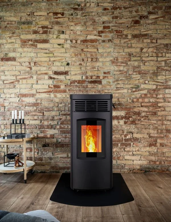Black freestanding pellet stove with glowing fire glass door on hearth mat against exposed brick wall